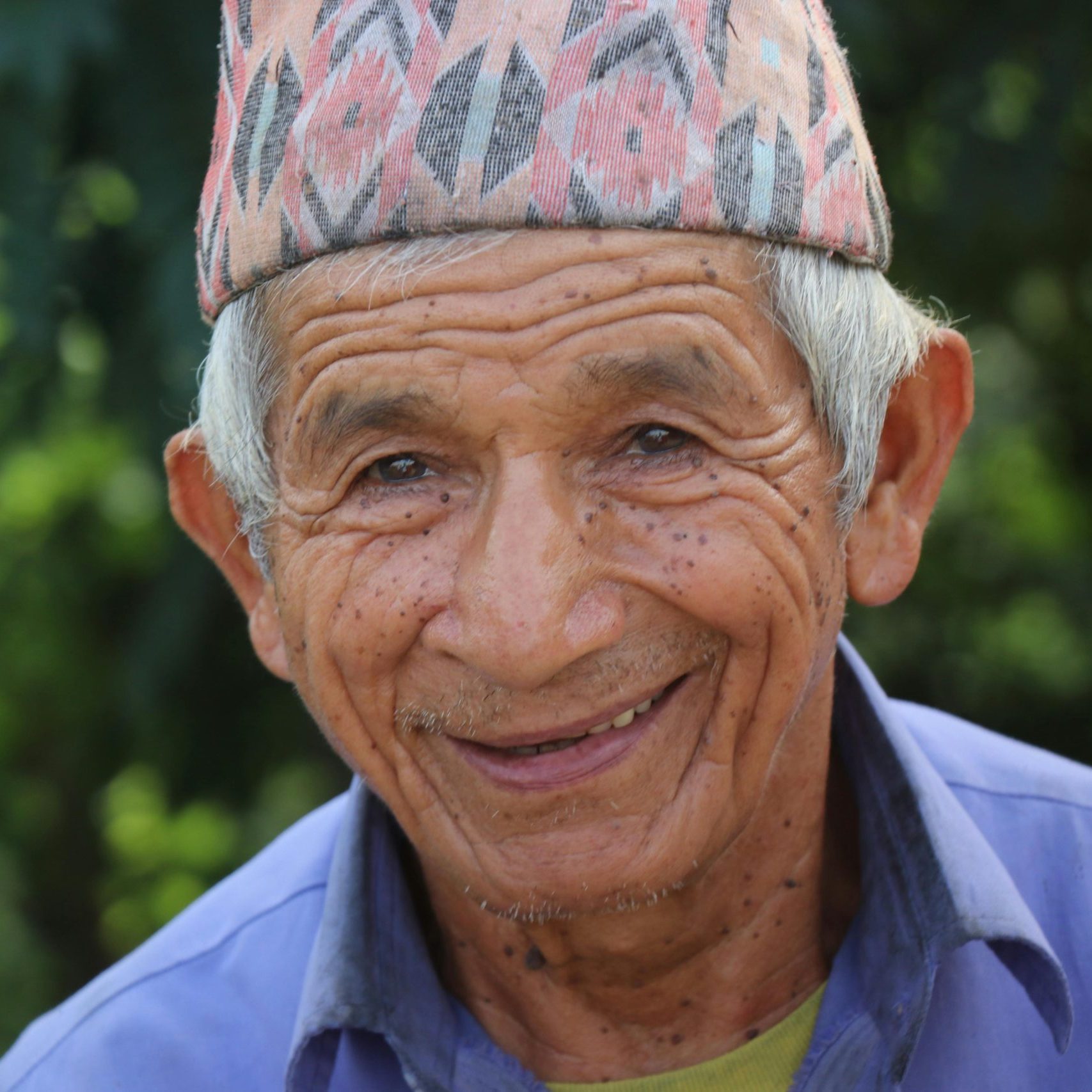 Portrait of an elderly man wearing a traditional dhaka topi, smiling warmly outdoors.
